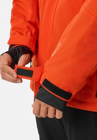 Giacca arancione con un esterno testurizzato, dotata di una fascia in Velcro al polsino e di una fodera interna nera per maggior calore e comfort.