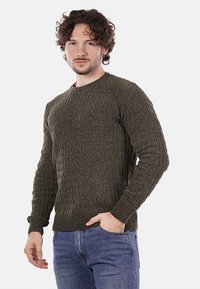 Jersey de punto verde oscuro con un patrón de cable texturizado, cuello redondo y puños y dobladillo acanalados, combinado con jeans azules.