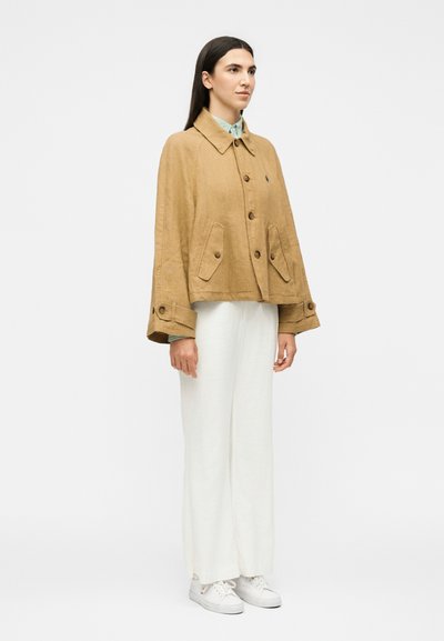 Γυναίκα όρθια με μπεζ κουμπωτό σακάκι, λευκό φαρδύ παντελόνι και λευκά αθλητικά παπούτσια, σε απλό φωτεινό φόντο.