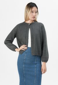 Žena v tmavě šedém silném pleteném kardiganu přes bílý top a sukni v vysokém pase z modrého denimu, pózující s rukou na boku.