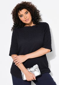 Schwarzes, strukturiertes Oberteil mit kurzen Ärmeln und gewelltem Ausschnitt, kombiniert mit einer silbernen Clutch. Fokus auf den Stoffdetails und dem entspannten Schnitt.