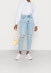 Jean bleu clair usé avec des déchirures aux genoux, associé à une chemise à boutons rayée blanche et bleue, et des baskets pastel. Sac à main jaune.