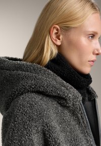 Jachetă din fleece gri cu glugă, având o textură moale și un sal din fleece negru pentru gât, prezentată pe profilul unui model, pe un fundal neutru.