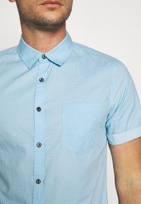 Himmelblaues Kurzarmhemd mit einem kleinen Musterdesign, Kentkragen und einer Brusttasche. Schwarze Knöpfe setzen einen Kontrast zum Stoff.