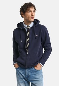 Navy zip-up hoodie with a drawstring, front pockets, and a logo on the chest, worn over a striped shirt and tie, paired with light blue jeans.