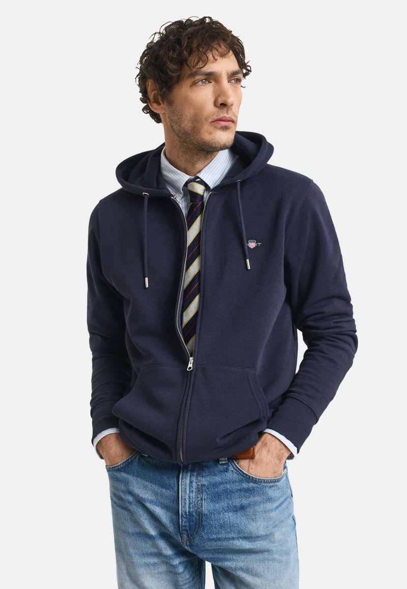 Navy zip-up hoodie with a drawstring, front pockets, and a logo on the chest, worn over a striped shirt and tie, paired with light blue jeans.