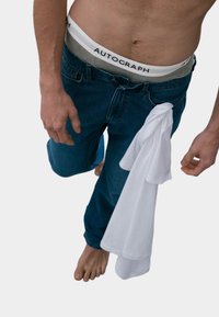 Jeans in denim blu scuro con un design a gamba dritta, tenuti in vita con una t-shirt di cotone bianca drappeggiata sopra. Vita grigia con il logo "AUTOGRAPH".