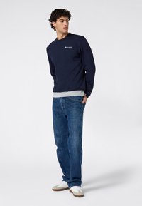 Sudadera azul marino con cuello redondo y logo bordado, combinada con una camiseta gris. Se lleva con vaqueros azul claro y zapatillas blancas.