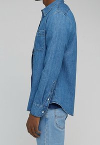 Persoon in een blauwe denim overhemd met knoopmanchetten en borstzak, gecombineerd met lichtblauwe denim jeans tegen een effen achtergrond.