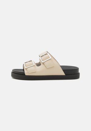 Beige liukuva sandaali, jossa on kaksi säädettävää hopeista solkea, musta kumipohja, sileä synteettinen päällinen ja minimalistinen muotoilu.