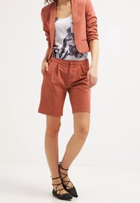 Lekka różowa marynarka zestawiona z kwiecistym topem na ramiączkach i dopasowanymi szortami. Materiał wydaje się gładki, z plisami na szortach oraz czarne buty na paski.