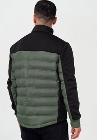 Chaqueta aislante en negro y verde olivo con textura acolchada en la parte trasera y tejido suave en el frontal. Cuenta con un collar tipo alto.