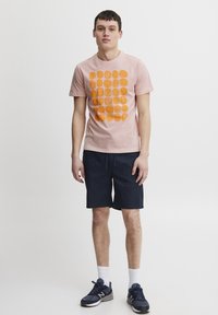 Lys rosa T-skjorte med oransje sirkulært mønster, kombinert med mørk marineblå shorts. Modellen har på seg hvite sokker og marineblå joggesko.