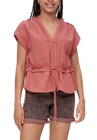 Femme portant un blouse en saumon texturée à manches courtes avec des boutons et une ceinture à la taille, assortie à un short en denim noir délavé avec des ourlets roulés.