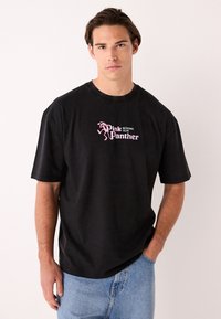 Črna bombažna majica s kratkimi rokavi in okroglim izrezom, ki prikazuje rožnato grafiko tekaškega Pink Pantherja in napis "Pink Panther Running Club".