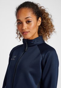 Marinblå idrottsjacka med hög krage, fullständig frontdragkedja och texturerad tyg. Har varumärkeslogga på vänster bröst och dragsnoddar som accent.