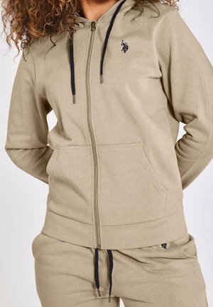 Personne portant un sweat à capuche beige zippé et un pantalon de survêtement assorti avec des cordons noirs et un petit logo brodé sur la poitrine et le pantalon.