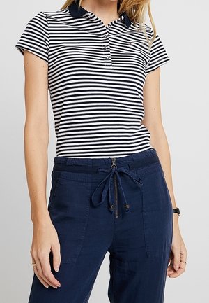 Vrouw die een navyblauw-wit horizontaal gestreept poloshirt met korte mouwen draagt en navyblauwe broek met trekkoord en voorzakken.