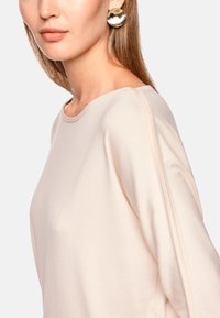 Light beige long-sleeve top with a round neckline and soft texture. The model wears large round gold earrings that add contrast.