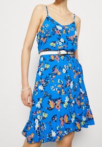 Sininen kukallinen mekko, jossa on ohuet h ihansuolet, istuva vartalo ja röyhelöinen helma. Mekko sisältää monivärisiä kukkia ja valkoisen vyön vyötäröllä.