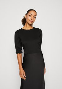 Black blouse with puffed sleeves and a round neck, made from textured fabric. The model is wearing a smooth black skirt, showcasing both garments.