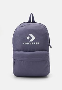 Mochila de tela morada con un bolsillo frontal, que presenta un logotipo y una estrella blanca de Converse. Incluye correas flexibles y un asa superior para llevar.