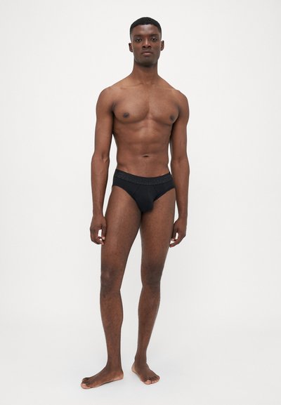 Schwarze Herrenunterhose mit einem strukturierten Bund, mit einer enganliegenden Passform und glattem Stoff, dargestellt an einem Modell vor einem schlichten Hintergrund.