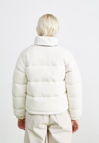 Bunda do deky v bielej fleece s vysokým golierom a viditeľným švom. Obsahuje elastické manžety a malé logo na golieri.