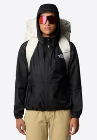 Black, lightweight hooded jacket with zipper; features elastic cuffs and hem, paired with a cream-colored backpack and reflective sunglasses.