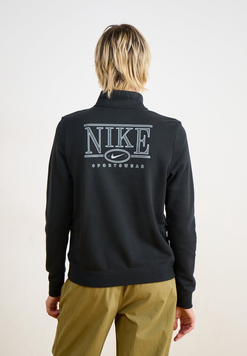 Svart Nike-hoodie med hög krage och stor vit text på baksidan. Ribbade ärmslut och nederkant. Modellen bär olivgröna byxor.