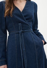 Mörkblå denimwrapklänning med långa ärmar, V-ringning, knytbälte i midjan och framfickor. Synlig sömnadsdetalj förhöjer designen.