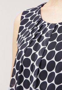 Top sin mangas en azul marino y blanco con un patrón de puntos, cuello plisado y textura suave. La tela parece ligera y fluida.