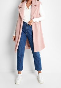 Long gilet rose clair doublé en polaire avec un col à revers encadré, porté sur un haut blanc, associé à un jean bleu taille haute et des baskets blanches.