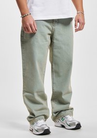 Jeans de mezclilla verde claro con un corte relajado, que presentan costuras visibles y una textura ligeramente desgastada, combinados con zapatillas plateadas y verdes.