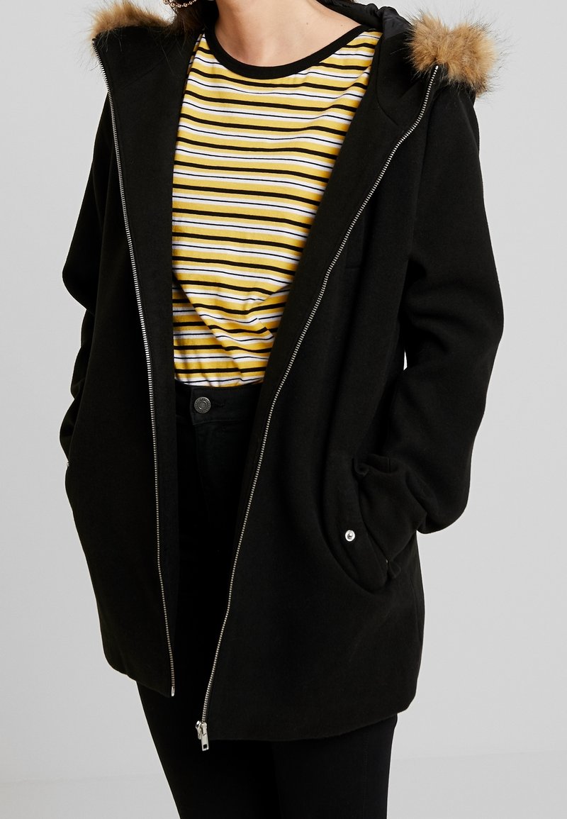 Manteau noir zippé avec capuche bordée de fourrure porté par-dessus une chemise rayée jaune, blanche et noire et un pantalon noir.