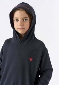 Giovane con capelli chiari e occhi azzurri che indossa una felpa con cappuccio di colore blu scuro con la scritta "U.S. Polo Assn." sul cappuccio e un logo rosso sul petto.