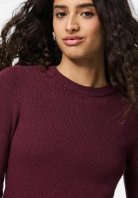 Burgundijas ribota adīta džemperis ar apaļu kakla izgriezumu un garām piedurknēm. Audumam ir gluda struktūra ar nelielu elastību.