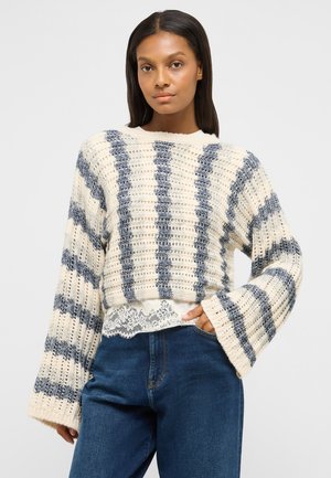 Woman wearing a cream and gray striped knit sweater with bell sleeves, white lace trim top, and blue jeans, standing with one hand in pocket.