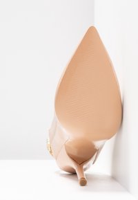 Chaussure à talon haut beige à bout pointu avec côtés transparents et talon fin, inclinée contre un coin blanc en arrière-plan.
