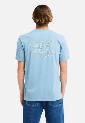 Junge Person mit kurzen braunen Haaren, die ein hellblaues T-Shirt mit einem Wellenmuster auf dem Rücken und blaue Jeans trägt.