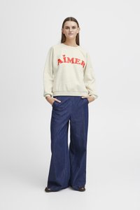 Cream sweatshirt with red "AIMER" text, paired with wide-leg dark blue denim trousers. Model stands with hands in pockets.