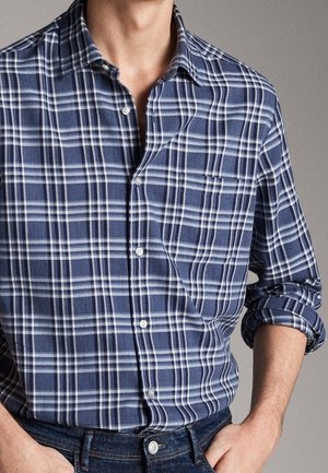 Homme portant une chemise à carreaux bleue et blanche, à boutons, avec les manches retroussées, et un jean bleu foncé.