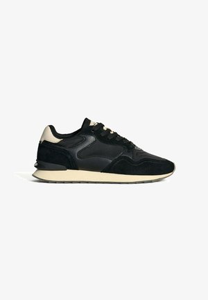 Zapatilla deportiva negra que combina materiales de ante y cuero, con una suela beige y detalles texturizados en los laterales. Punta redondeada y cordones.