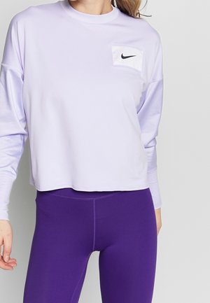 Femme portant un haut court Nike à manches longues de couleur violet clair et un legging taille haute violet foncé, debout devant un fond gris.