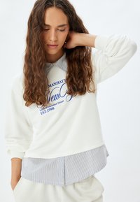 Sudadera blanca con un estampado gráfico azul. Presenta un dobladillo de camisa de cuello a rayas, mangas largas y un corte relajado. Textura de tela suave.