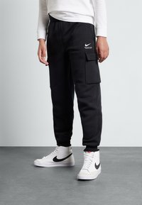 Schwarze Cargo-Hosen aus weichem Material mit elastischem Bund. Mit großen Seitentaschen und einem weißen Nike-Logo. Kombiniert mit weißen High-Top-Sneakern.