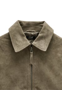 Zamszowa kurtka w stonowanej oliwkowej zieleni, z wyrazistym kołnierzem i zamkiem błyskawicznym z przodu. Miękka faktura z minimalnymi detalami przeszycia. Widoczna metka marki.