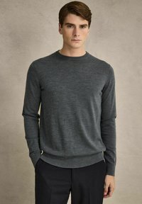 Grå långärmad tröja i mjukt tyg med rund halsringning, med en figurnära midja och ärmmuddar, bärs över mörka byxor.