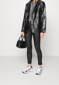 Person som bär en glänsande svart texturerad kavaj, metallisk silverfärgad topp, svarta åtsittande byxor, vita sneakers och håller en liten svart handväska med två handtag.