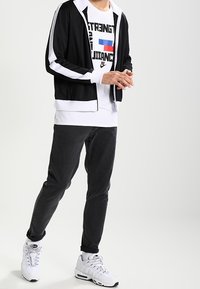 Black zip-up jacket with white stripes, white graphic t-shirt with text, dark gray jeans, and white sneakers.